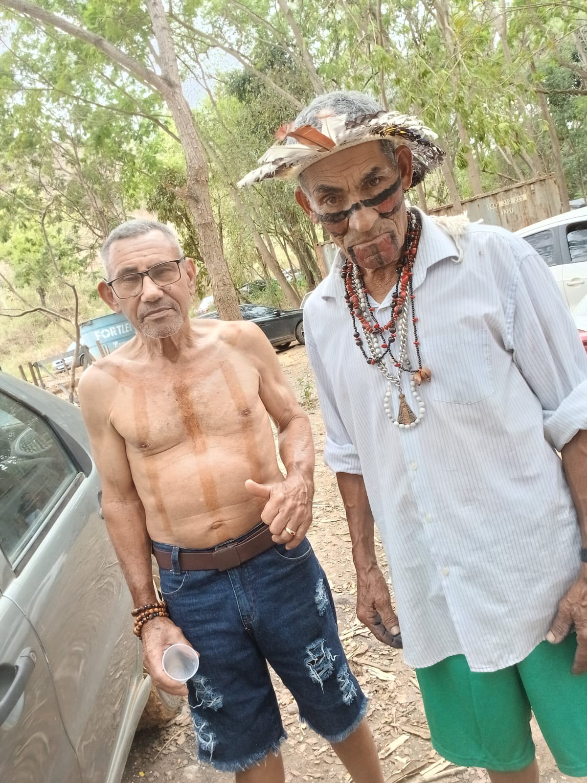 Dois homens juntos ao ar livre, um deles com cocar, colares e pintura no rosto